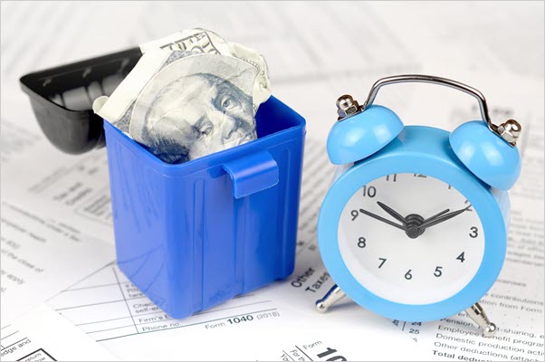 Toy garbage can containing a crumbled $10 bill next to a toy alarm clock, and both are sitting on a pile of budget documents.