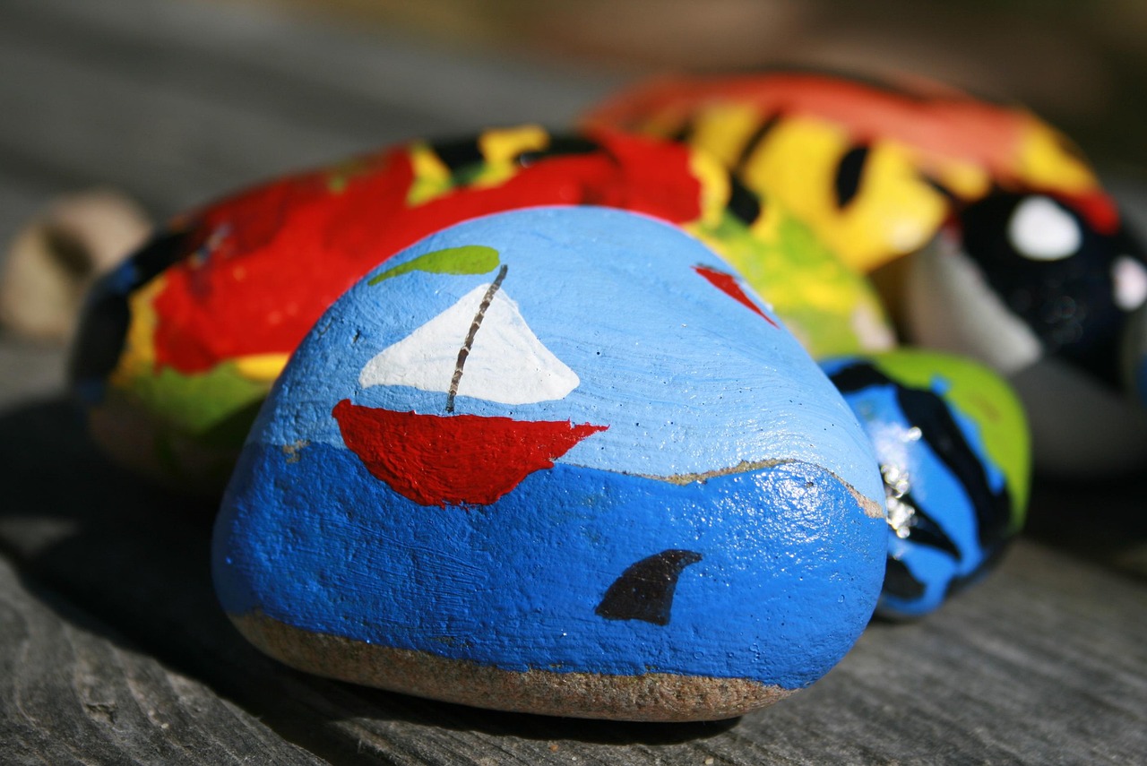 Painted rocks on a wooden surface, featuring a blue rock with a sailboat and shark fin design.