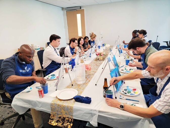 People sitting at long tables engaged in a painting activity, focused on individual canvases. Several painting supplies are scattered on the tables.