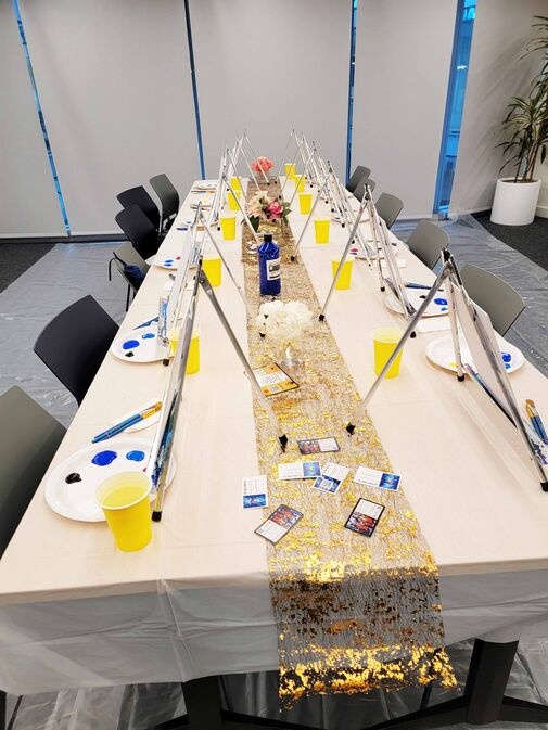 A long table set up for a painting activity with easels, canvases, paint supplies, and yellow cups. A gold sequin runner decorates the center, with a few flowers.