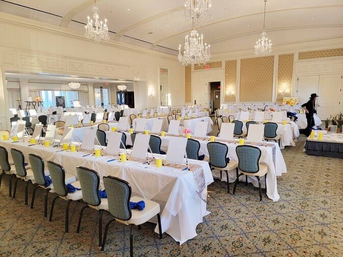 A banquet hall with rows of tables and chairs set for an event. Tables are covered with white tablecloths and have yellow cups placed on them. Chandeliers hang from the ceiling.