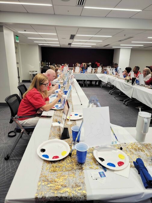 People sitting at a long table participating in a painting class, with canvas, paint, and brushes in front of them.